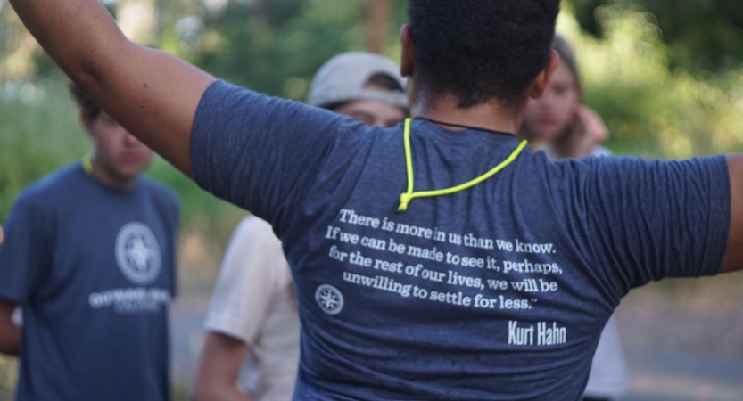 A person wearing an outward bound shirt lifts their hands in the air. The back of the shirt reads, "There is more in us than we know. If we can be made to see it, perhaps for the rest of our lives, we will be unwilling to settle for less. – Kurt Hahn"